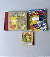 Photo of three Simpsons collectible books: The Simpsons Handbook, The Simpsons Holiday Humdinger, and The Bart Book on a plain surface.