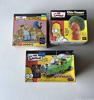 Brand new unopened Simpsons collectibles lot including puzzle, Chia Homer planter, and snap-together car kit. Boxes show wear.