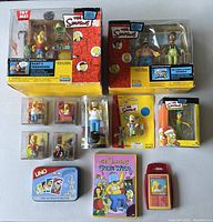 Photo showing unopened interactive diorama boxes of Bart's Treehouse and Kwik-E-Mart, along with small boxed and loose collectibles including figures, keychain, and card games.