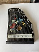 Photo of a Jubel Tone Konzert-Salon-Harfe Zither showing black wood with colorful floral painting, sound hole, strings, tuning pins, and tuning key chart attached.