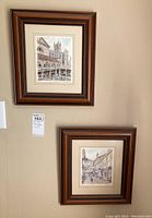 Two framed artworks hanging on a wall showing city scenes in wooden frames.
