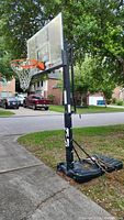 Full view of the basketball hoop set up outdoors near a sidewalk and driveway. Hoop and backboard in clear view with base filled and stabilized.