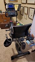 Photo showing the full Schwinn exercise bike from the rear, featuring black ventilated bicycle-style seat with backrest, digital display console at the front, a cup holder on the side, and the bike frame and pedals.