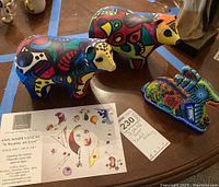 Photo shows two brightly hand painted bear figurines with abstract multicolor designs and one beaded donkey figurine with some beads missing, placed on a wooden surface with a postcard and a sale tag.