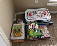 Photo of SureShot table hockey game with pieces on ice rink, stacked board game boxes including Monopoly and Last Word, several boxed board games and nature-themed game.
