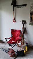 Photo showing a red foldable chair, two shovels, a red electric leaf blower, and several plastic garden fluid containers.
