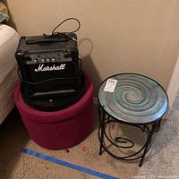 Photo showing the Marshall MG10CF guitar amplifier on top of red fabric ottoman, placed next to a round metal table with spiral glass top