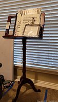 Wooden music stand with carved central column and tripod base, holding multiple sheets of music and a small plastic container.
