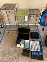 Three metal plant stand side tables displayed together with multiple jewelry boxes, eyeglass cases, and folded cloth pieces arranged below them on a carpeted floor.
