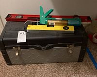 Side view of closed black and silver tool box with metal latches, red level, yellow ruler, and green manual caulking gun dispenser on top.