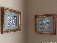 Two gold colour framed wall art pieces hanging on a wall, one with a cafe scene and one with a country scene