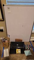 Full view showing whiteboard, HP printer, markers, eraser, small card magnets, portfolio, books, and small brown bag on carpet.
