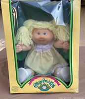 Front view of vintage Cabbage Patch Kids doll in yellow dress, yarn hair pigtails, inside yellow box with clear plastic front.
