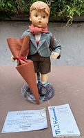 Front view of M.J. Hummel Little Scholar doll holding umbrella with certificates at front base