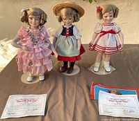 Three Shirley Temple Danbury Mint collectible dolls standing side-by-side on table with two certificate cards laid out.