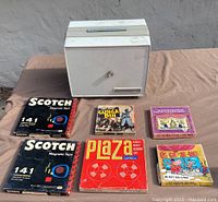 Vintage magnetic tapes and film reels displayed beside a white box container. Includes Scotch 141 tape packs, Plaza tape reel, and 8mm films 'Gunga Din', 'Les Crescendos', and 'Feet of Winds'.