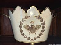 Photo of cream-colored ceramic cachepot decorated with a bee surrounded by a laurel wreath, scalloped rim, gold handles and feet