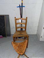 Front view of wooden ladder-back chair with rattan seat, brown leather tripod stool with carved decorations, and mop handles behind them against a white basement wall.