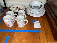 Photo showing coffee mugs with black script pattern and stacked bowls and plates with black rims and script-stamp designs.