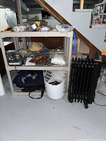 Full shelf contents with assorted items including VHS tapes, cords, painting supplies, laundry basket, and folded fabric or clothing under a staircase in a basement.