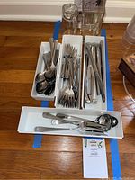 Overall view of stainless steel flatware organized by type in white trays along with two tall clear glass vases behind trays on wood floor.