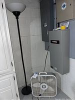 Black floor lamp and white box fan placed in a basement corner next to electrical panels.