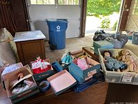 Overall view showing nine Madame Alexander Little Women dolls in boxes and several other vintage dolls arranged on a table and in plastic storage bins in a garage setting.