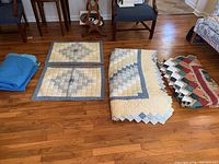 Photo showing two patchwork quilts with scalloped edges in blue, beige, and cream tones, two matching sham pillows, and a folded solid blue blanket on hardwood floor.