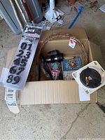 View of entire lot in garage including reflective numbers in packaging, grapevine wreath, black travel mug, disposable coffee cup, jar opener, weatherseal spool, and other small items in cardboard box.