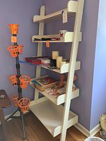 Wide shot of cream colored wooden shelf unit with five shelves, showing assortment of candles, mugs, boxes, and a doll on the shelves. Basketball game stand to the left with multiple orange hoops attached.