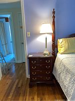 Dark wooden night stand next to bed with white quilt and yellow pillow. Lamp is turned on on top of the night stand.