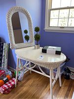 Front view of white wicker table with arched wicker mirror, two small faux greenery topiary plants, and boxed zen garden on tabletop