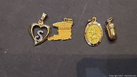 Four 10k gold charms displayed on a dark background: heart with 'S', Trinidad map, religious medal, small capsule.