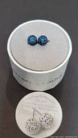 Close-up of two blue crystal ball stud earrings displayed on a light grey circular stand with Helberg & Burke branding.