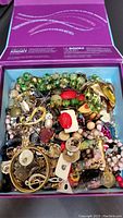 Photo showing a variety of beaded necklaces (green and beige beads), rings with large stones, gold bangles, and brooches in an open box.