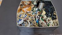 Metal box filled with assorted mixed costume jewelry pieces including various bracelets, necklaces, beads, and a metal thimble.