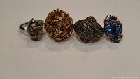 Four assorted rings with various metal tones and stone settings photographed together on a flat surface.
