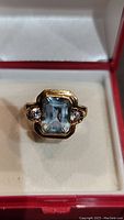 Front view in red and white box showing emerald-cut aquamarine with two round white accent stones