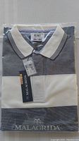 Photo of folded men's Malagrida polo shirt in original plastic packaging showing color block design, white collar with navy stripes, tags attached, and embroidered logo on front.