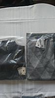 Pair of men's casual shirts folded on a white surface, one light grey checkered sweater and one charcoal shirt with ace card graphic, both in clear plastic bags.
