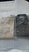 Both men's tops, sand sweater on left and grey button shirt on right, in original packaging on white surface.