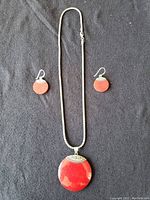 Photo showing the full set with necklace and earrings on black fabric background. Necklace has a large red pendant with silver detail, accompanied by matching red circular earrings with silver detail.