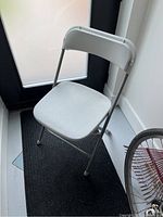 Off white folding chair made of plastic and metal, shown standing on black mat near door.