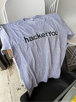 Light gray HackerYou t-shirt with black logo on front laid on chair showing front design.