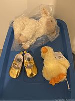 Photo showing all three items together on a blue plastic bin: white sheep stuffed animal sealed in plastic, yellow baby duck with tags, and gold baby shoes.