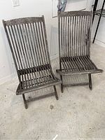 Pair of two wooden patio folding chairs with a weathered gray finish shown side by side against a white wall and concrete floor.