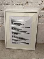 Front view of framed print showing Morse code alphabet layout in black on white inside white frame standing against wall.