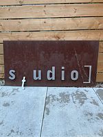 Wide shot showing full rectangular metal sign with letters spelling 'studio'. The 'T' letter is detached and laying below the sign.