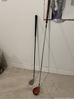 Two golf clubs shown standing against a wall; left club with dark grip and silver head, right club with orange head and silver shaft.