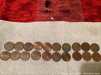 Full view of 20 Canadian pennies in two rows on white cloth with '1980-1999' written on plastic bag in background, showing wear and oxidation on coins.